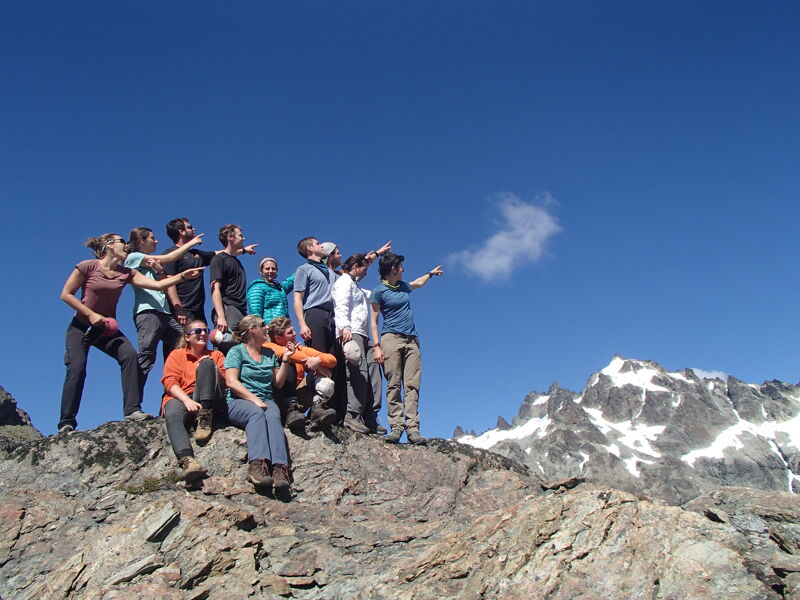 A group of about a dozen people are standing and sitting on a rocky outcrop, possibly a mountain summit, under a clear blue sky. They appear to be looking towards the right side of the frame, possibly at a mountain range partially covered with snow. The overall impression is one of camaraderie and enjoying a scenic view after a hike.